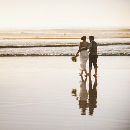 Bride and groom walk barefoot along wet sand at sunset, reflected in the shallow water